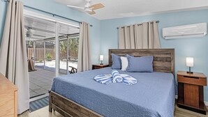 Une chambre à coucher comprenant un lit, une table de chevet, un ventilateur de plafond, une fenêtre avec des rideaux et un appareil de climatisation.