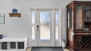 Une porte d’entrée avec des panneaux de verre dépoli, un banc blanc doté de compartiments de rangement, un meuble en bois avec des portes en verre et un paillasson gris.