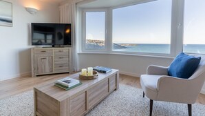 Un salon moderne doté d’une grande fenêtre donnant sur la côte, d’un téléviseur à écran plat posé sur un meuble en bois et d’un fauteuil confortable.