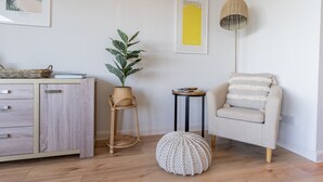 Une pièce chaleureuse avec un bureau en bois, une chaise en osier, un pouf rond, un fauteuil rembourré et une petite table d’appoint avec une lampe.