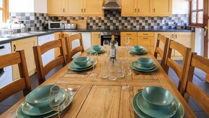 On aperçoit un coin repas avec une table en bois dressée pour six personnes, entourée de chaises en bois. Au fond, on distingue une cuisine avec un dosseret à carreaux et des placards en bois.