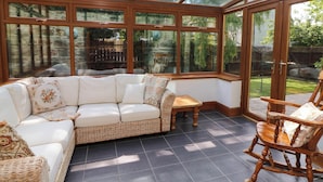 Une véranda avec un canapé d’angle blanc, des meubles en osier et un fauteuil à bascule en bois.