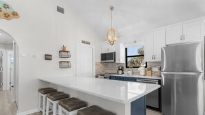 A modern kitchen with white cabinets, a stainless steel refrigerator, and a central island with bar stools. - 130 Tiki Tides: 2br W/ Outdoor Tiki Bar! 2 Bedroom Home (Fort Myers Beach)