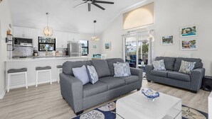 A modern living room with a grey sofa, white bar stools, a white coffee table, and a ceiling fan. - 130 Tiki Tides: 2br W/ Outdoor Tiki Bar! 2 Bedroom Home (Fort Myers Beach)
