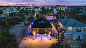 A beachfront property with a well-lit house, a paved driveway, and surrounding palm trees. - 130 Tiki Tides: 2br W/ Outdoor Tiki Bar! 2 Bedroom Home (Fort Myers Beach)