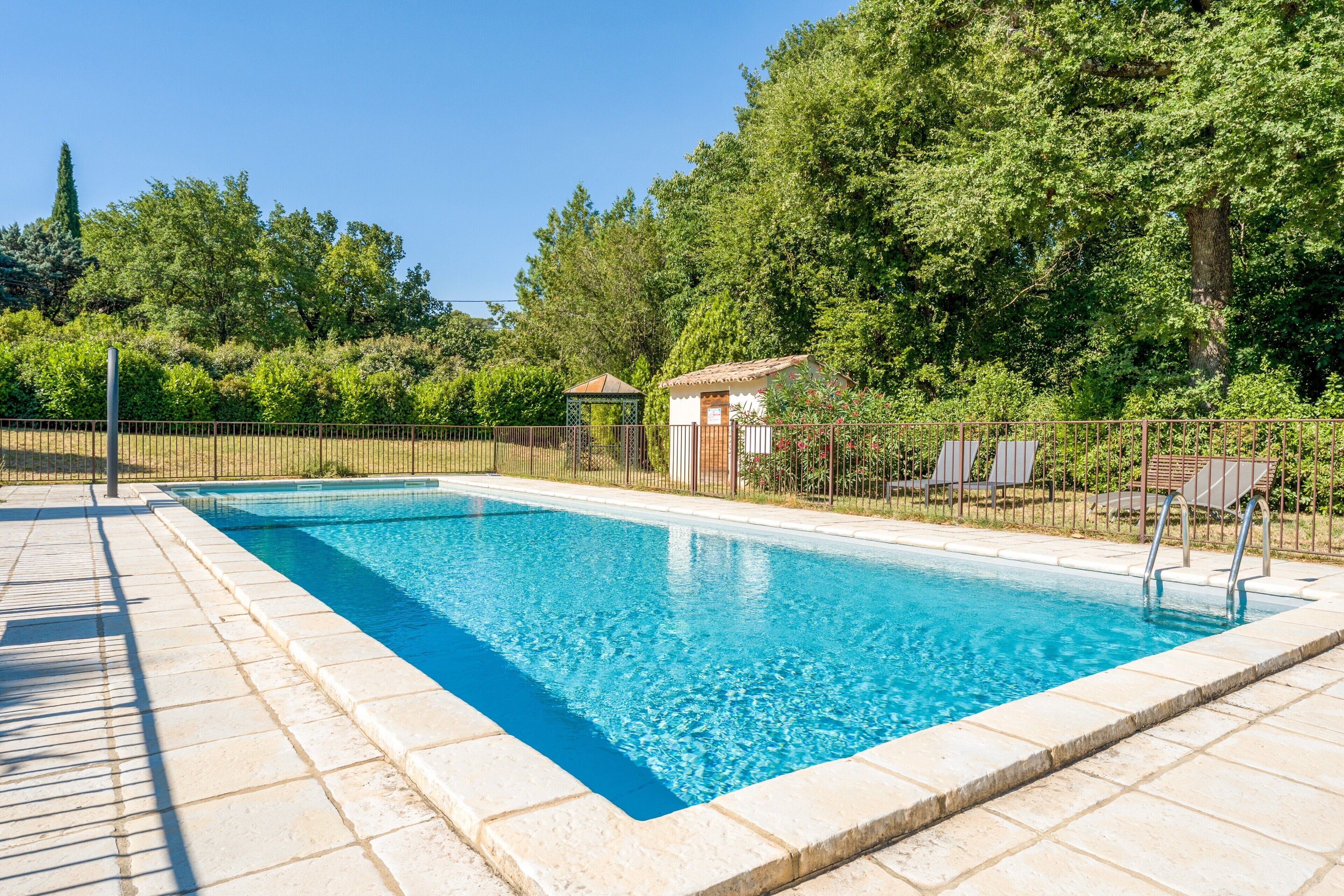 A rectangular swimming pool with a tiled edge, surrounded by a fence, and a small gazebo-like structure in the background.