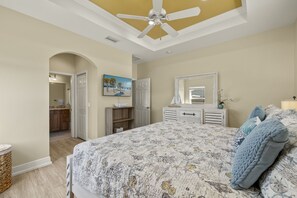 A bedroom with a large bed, a ceiling fan, a dresser, a mirror, and a door leading to a bathroom.