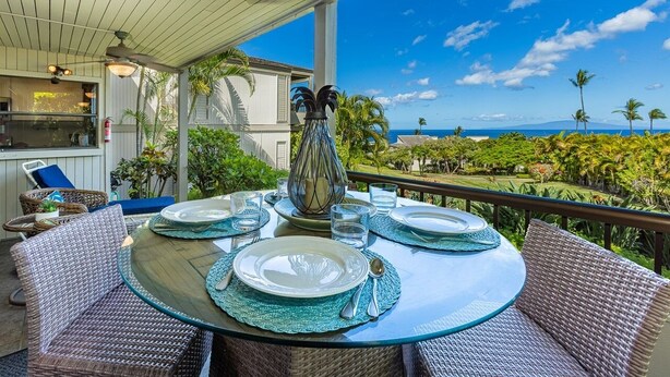 A round glass table set for four with plates, cutlery, and glasses, surrounded by wicker chairs, overlooking a scenic view of a lush landscape and ocean. - Wailea Ekolu 803 1 Bedroom Condo (Kihei)