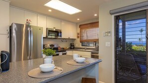 A modern kitchen with stainless steel appliances, a central island with bowls, and a sliding glass door to a balcony. - Wailea Ekolu 803 1 Bedroom Condo (Kihei)