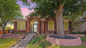 A two-story brick house with a front porch, a large tree in the front yard, and a well-maintained lawn.