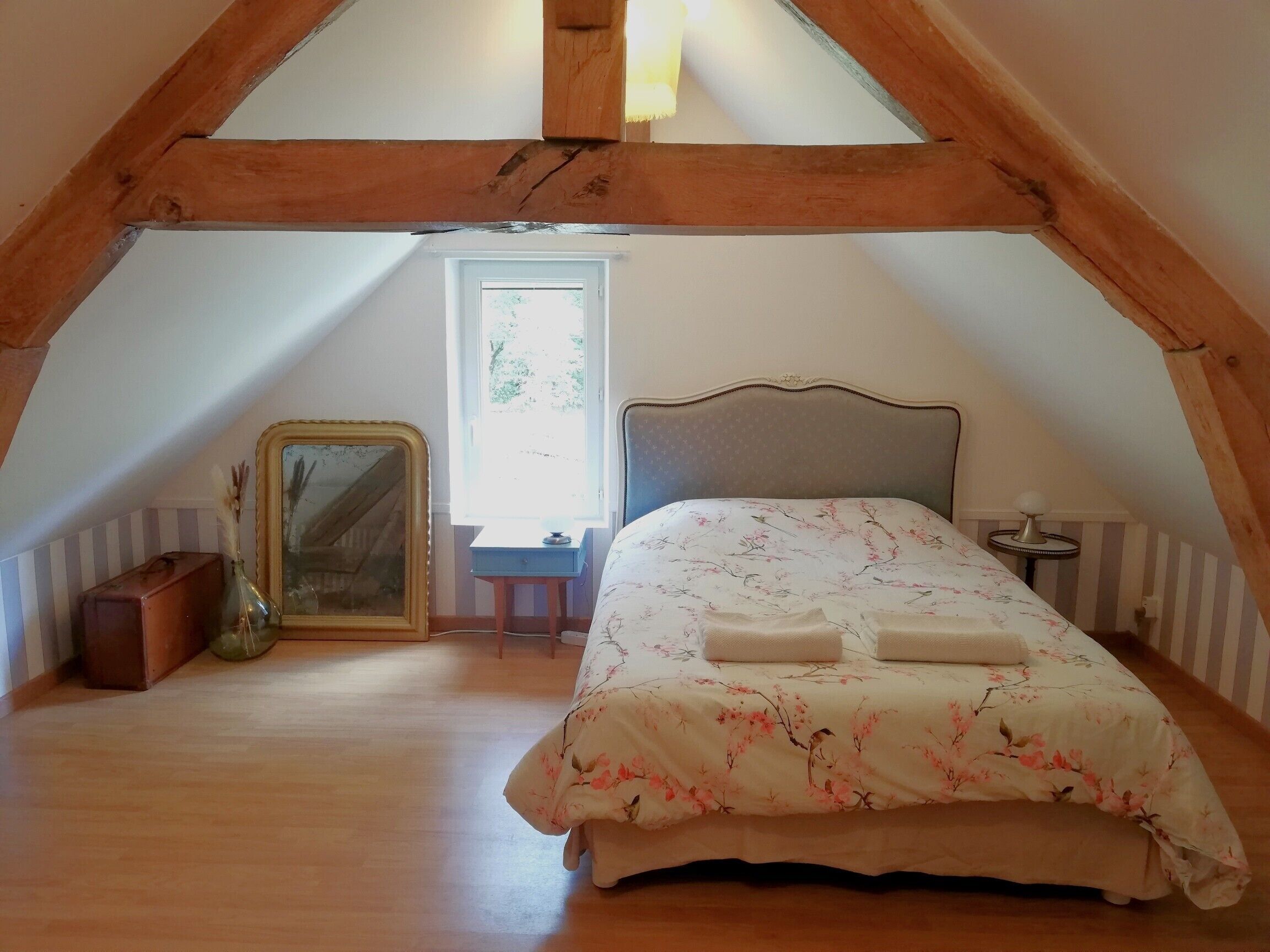 Une chambre avec un plafond en pente, un lit avec une literie à motifs floraux, une table de chevet, une lampe et un miroir.