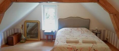 Une chambre avec un plafond en pente, un lit avec une literie à motifs floraux, une table de chevet, une lampe et un miroir.