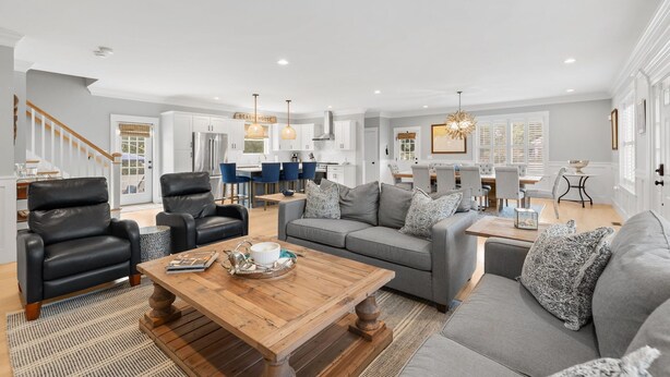 A spacious living room with a grey sofa, black leather armchairs, and a wooden coffee table. A kitchen with white cabinets and blue chairs is visible in the background.
