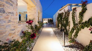 Un étroit sentier bordé de murs de pierre et de murs blancs recouverts de plantes grimpantes, qui mène à un bâtiment doté d’un toit en bois.