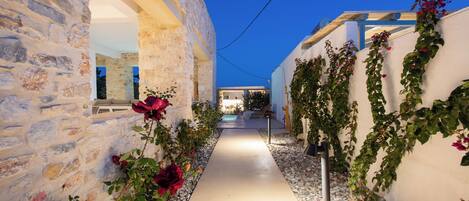 A narrow pathway flanked by stone walls and white walls with climbing plants, leading to a building with a wooden roof.