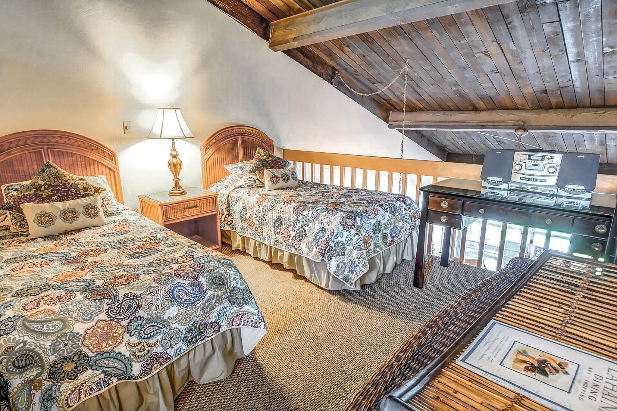 Ein Schlafzimmer mit zwei Betten, einem Holz-Kopfteil, einem Nachttisch mit Lampe sowie einem Glaskasten mit Audioanlage.
