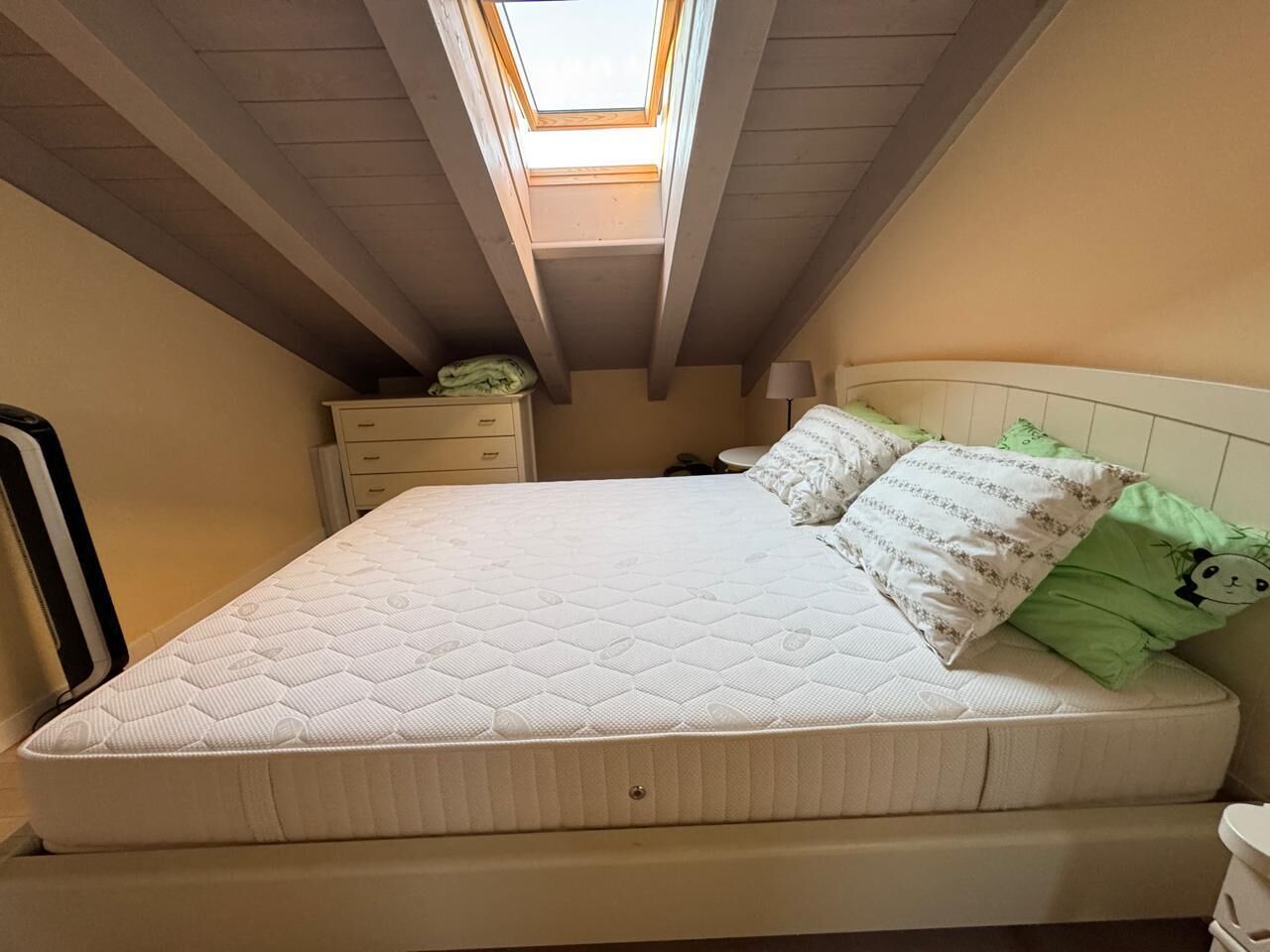 Une chambre avec un puits de lumière, un lit avec une couette, une table de chevet et une commode.