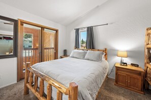 Un dormitorio con estructura de cama de madera, una mesita de noche con lámpara y vista al exterior a través de una puerta.