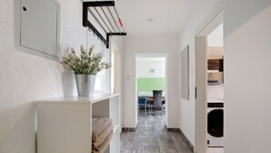 Un couloir avec une étagère blanche, une plante en pot, un porte-manteau suspendu et une vue sur une pièce où se trouve un lave-linge.