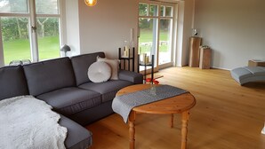 A modern living room with a sectional sofa, a wooden coffee table, and large windows offering a view of a garden. - Holiday Home 'Schäferhaus' with Private Terrace, Shared Garden and Wi-Fi (Hemme)