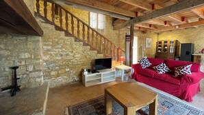 Un interior rústico con vigas de madera a la vista, pared de piedra, sofá rojo con cojines estampados, mesa de centro de madera y una escalera.