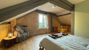 Un dormitorio acogedor en el ático, con techo inclinado, una cama grande, una mesita con lámpara, un sofá y una ventana con cortinas.