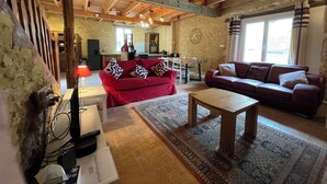 Una sala de estar acogedora con un sofá rojo, una mesa de centro de madera y una alfombra estampada. Se ve una cocina al fondo con paredes de piedra y vigas de madera.