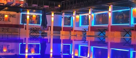 Zona de piscina con luces de neón azules y una zona de bar con un letrero de neón.