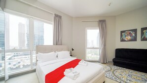 A modern hotel room with a large bed, a black sofa, and a city view through large windows. - PVH - Sunny Studio - Madison Residency (Dubai)