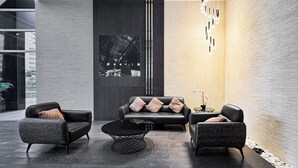 A modern lobby with a black leather sofa, two armchairs, a coffee table, and a small table with a plant. A large abstract artwork is on the wall, and there is a vertical light fixture. - PVH - Sunny Studio - Madison Residency (Dubai)