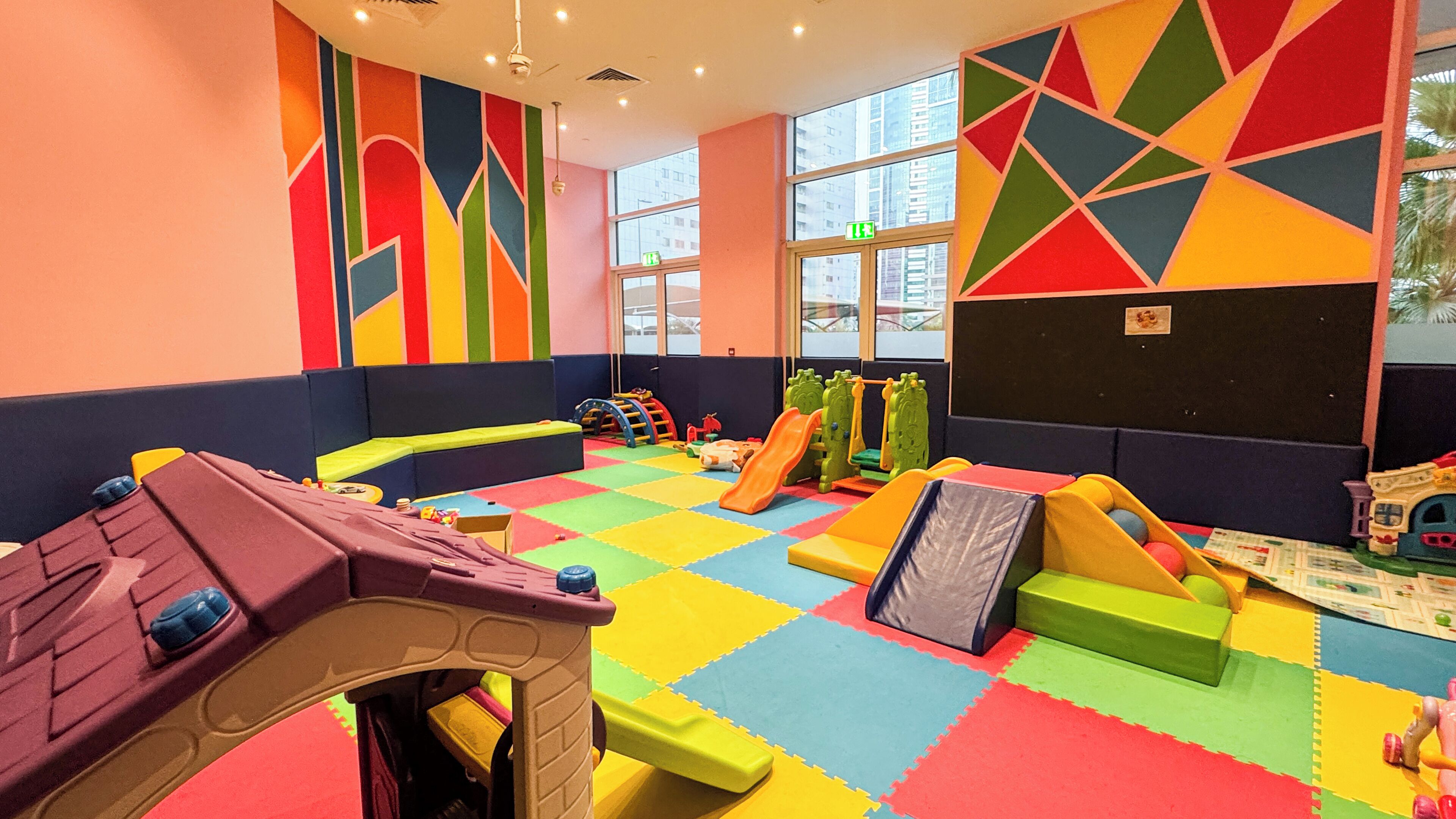 A children's play area with colorful foam mats, play structures, and a large window providing natural light.
