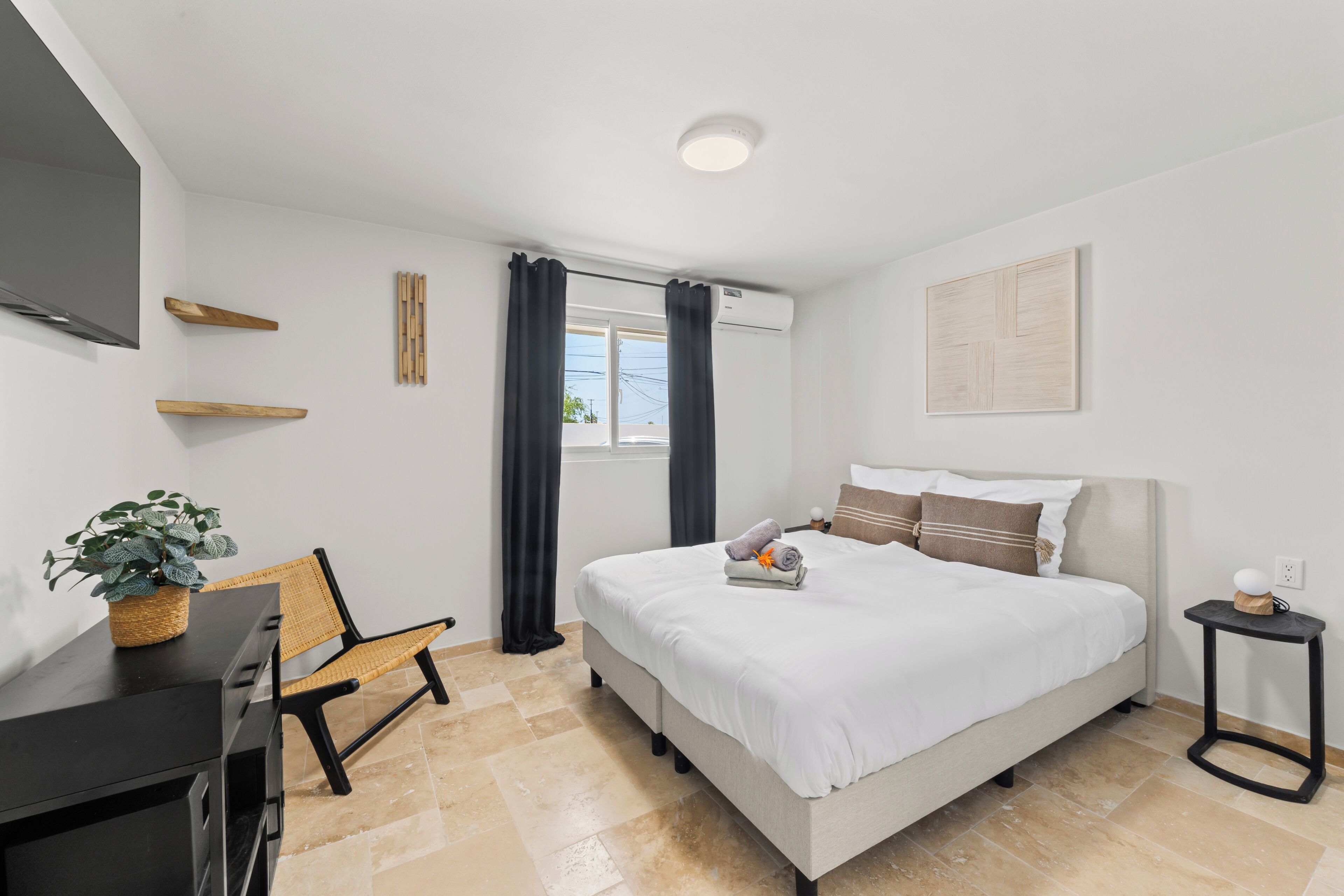 A modern hotel room with a bed, a desk, a chair, a potted plant, and a window with curtains.