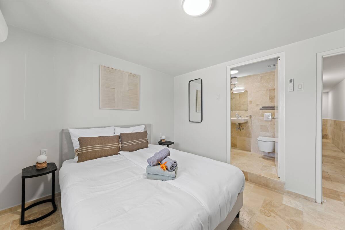 A hotel room with a neatly made bed, a small bedside table with a lamp, and a bathroom visible through an open door.
