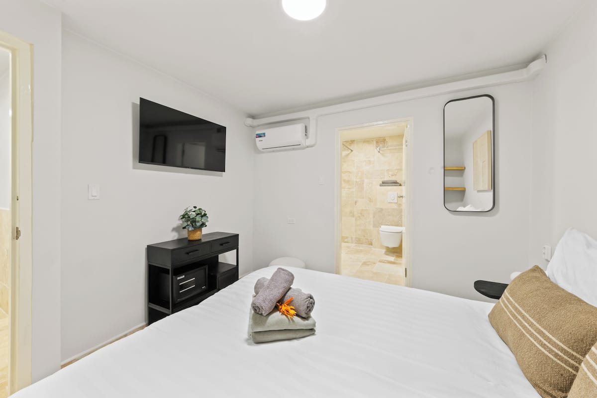 A hotel room with a bed, a small desk with drawers, a potted plant, a wall-mounted air conditioner, a mirror, and a bathroom door.