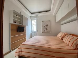 A bedroom with a bed, a TV, a window, and a wall-mounted shelf.