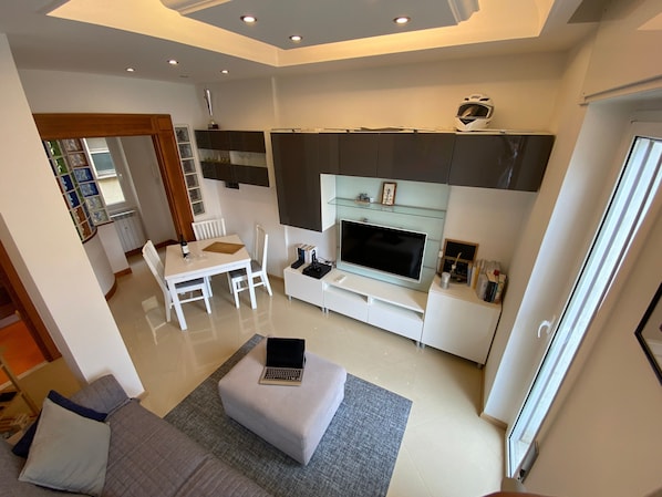 A modern living room with a flat-screen TV, a sofa, a dining table with chairs, and a laptop on a ottoman.