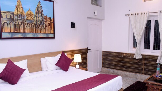 Habitación de hotel con una cama grande, una mesita de noche con lámpara, una ventana con cortinas y un cuadro de un edificio en la pared.