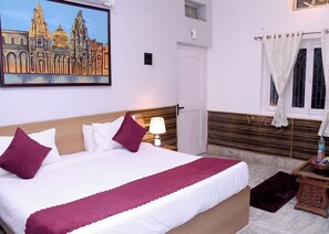 Quarto de hotel com uma cama grande, uma mesa de cabeceira com luminária, uma janela com cortinas e um quadro de um edifício na parede.