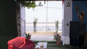 Habitación con una silla roja, una pequeña mesa y un televisor sobre un mueble bajo. Se ve un río y una barca al fondo.