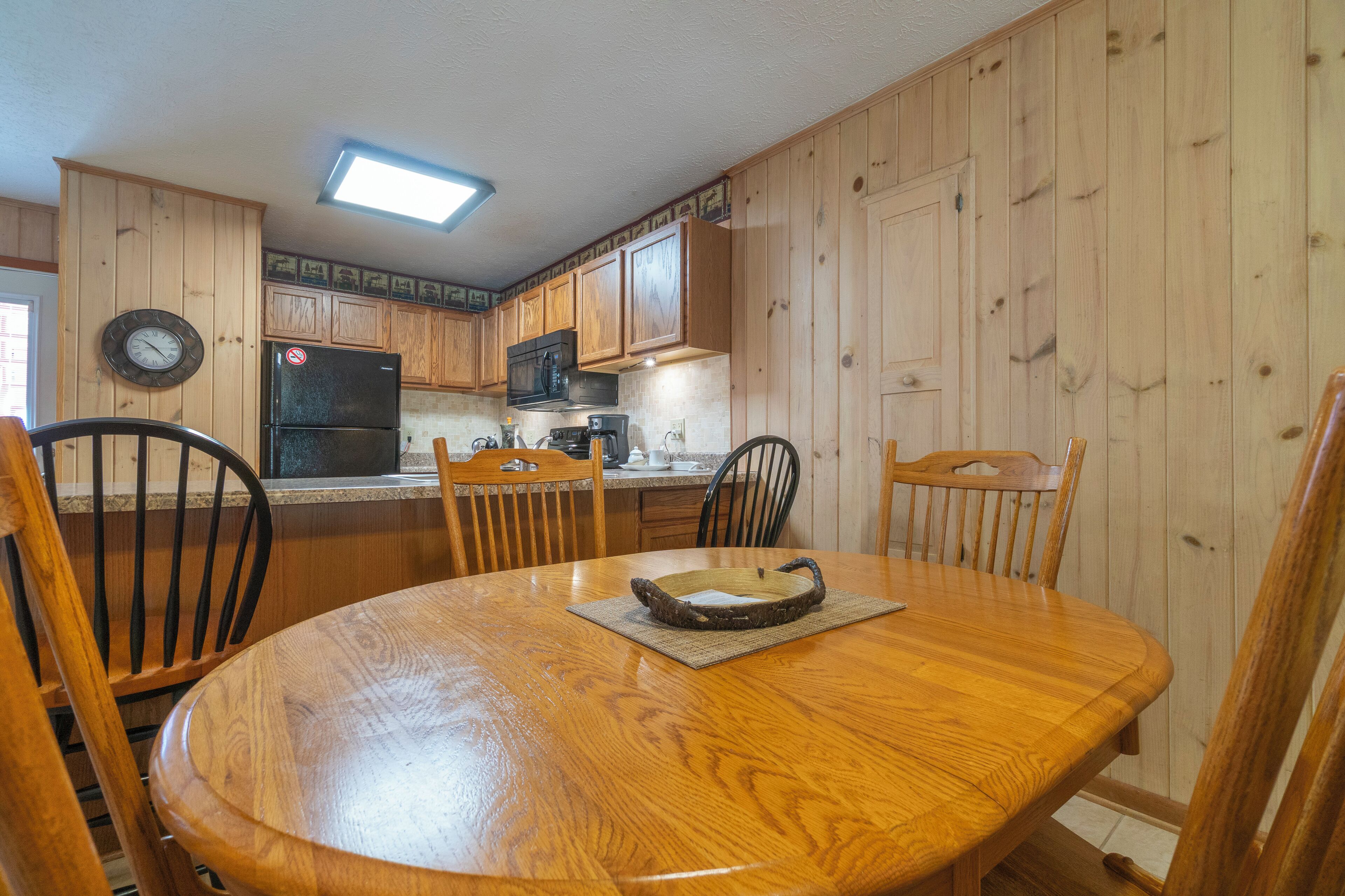 A wooden-paneled kitchen with a round dining table and chairs, a refrigerator, microwave, and a clock on the wall.