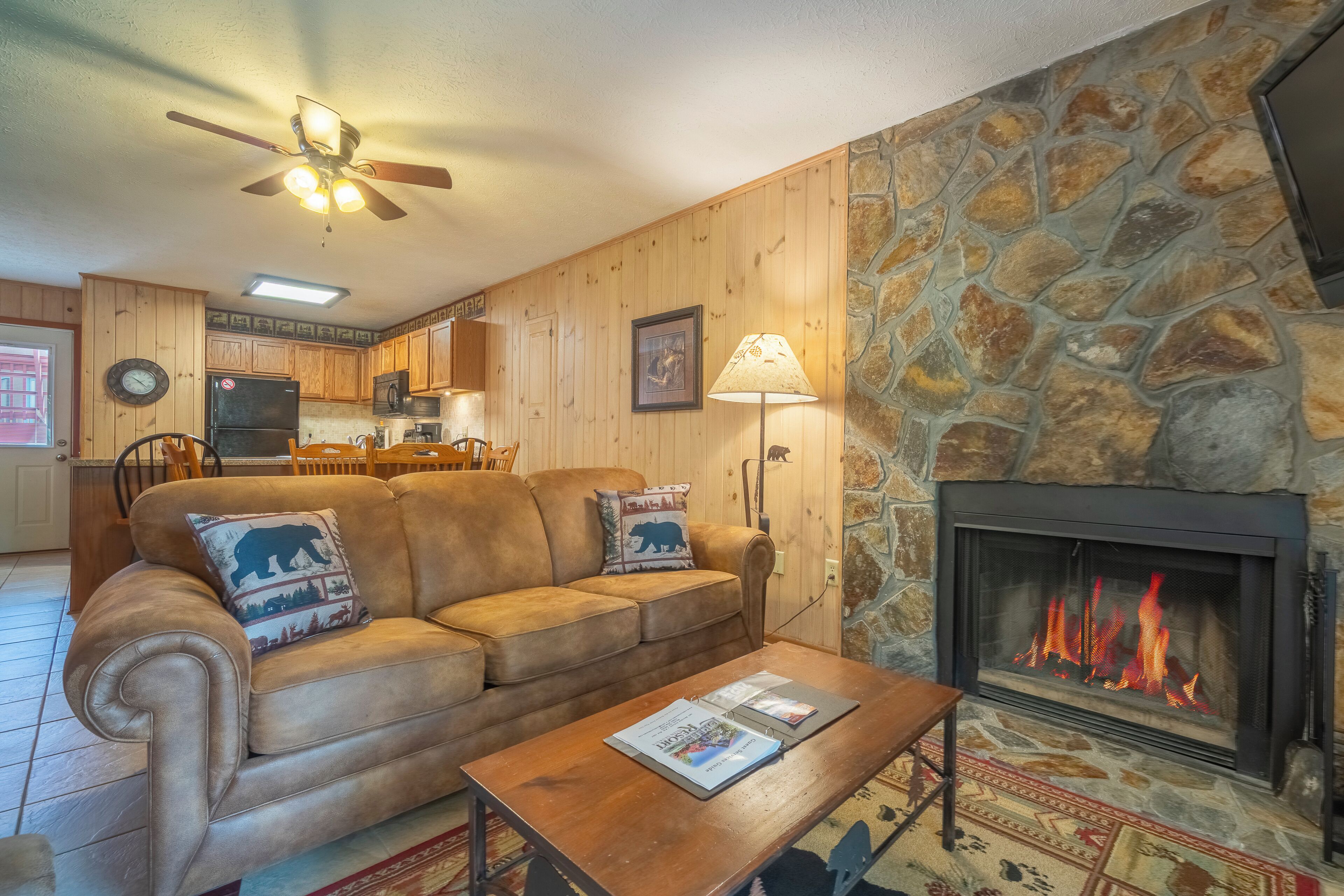 A cozy living room with a stone fireplace, a leather sofa with bear-themed pillows, a wooden coffee table, and a ceiling fan.