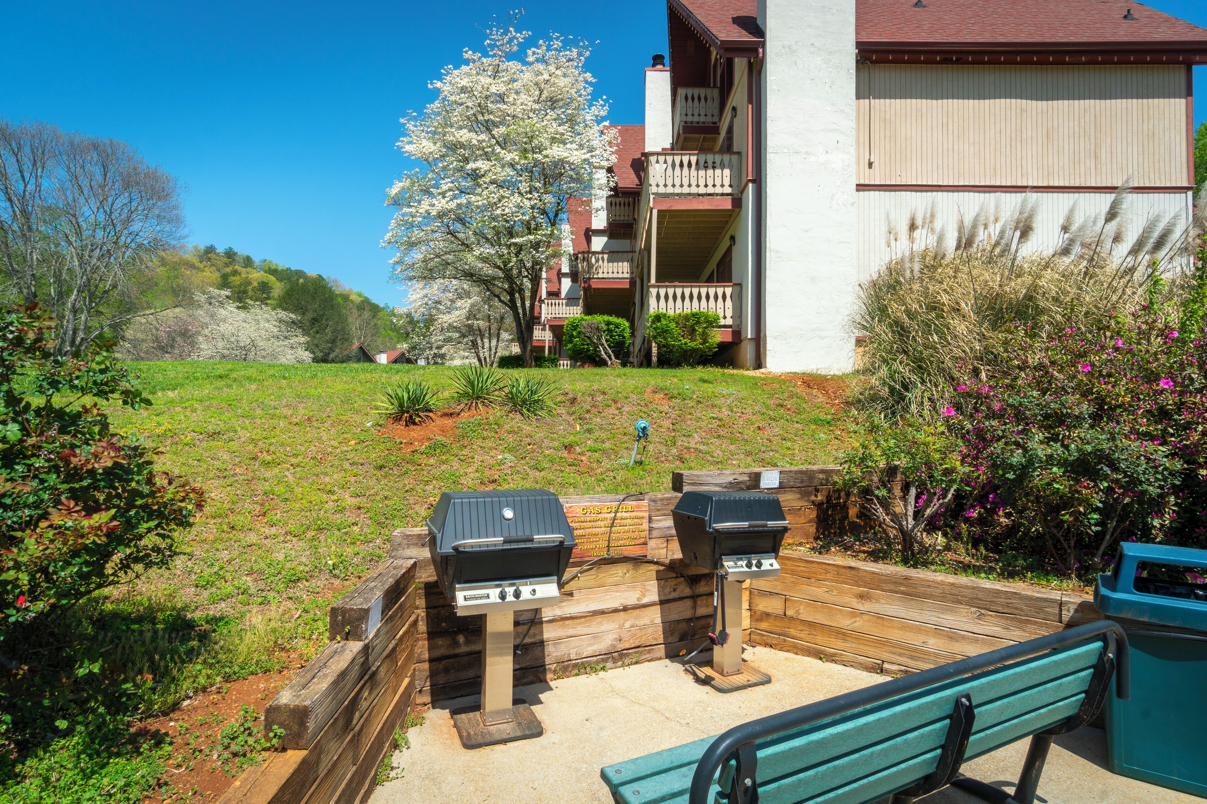 A patio area with two barbecue grills, a wooden bench, and a view of a residential building with a balcony.
