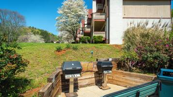 A patio area with two barbecue grills, a wooden bench, and a view of a residential building with a balcony.