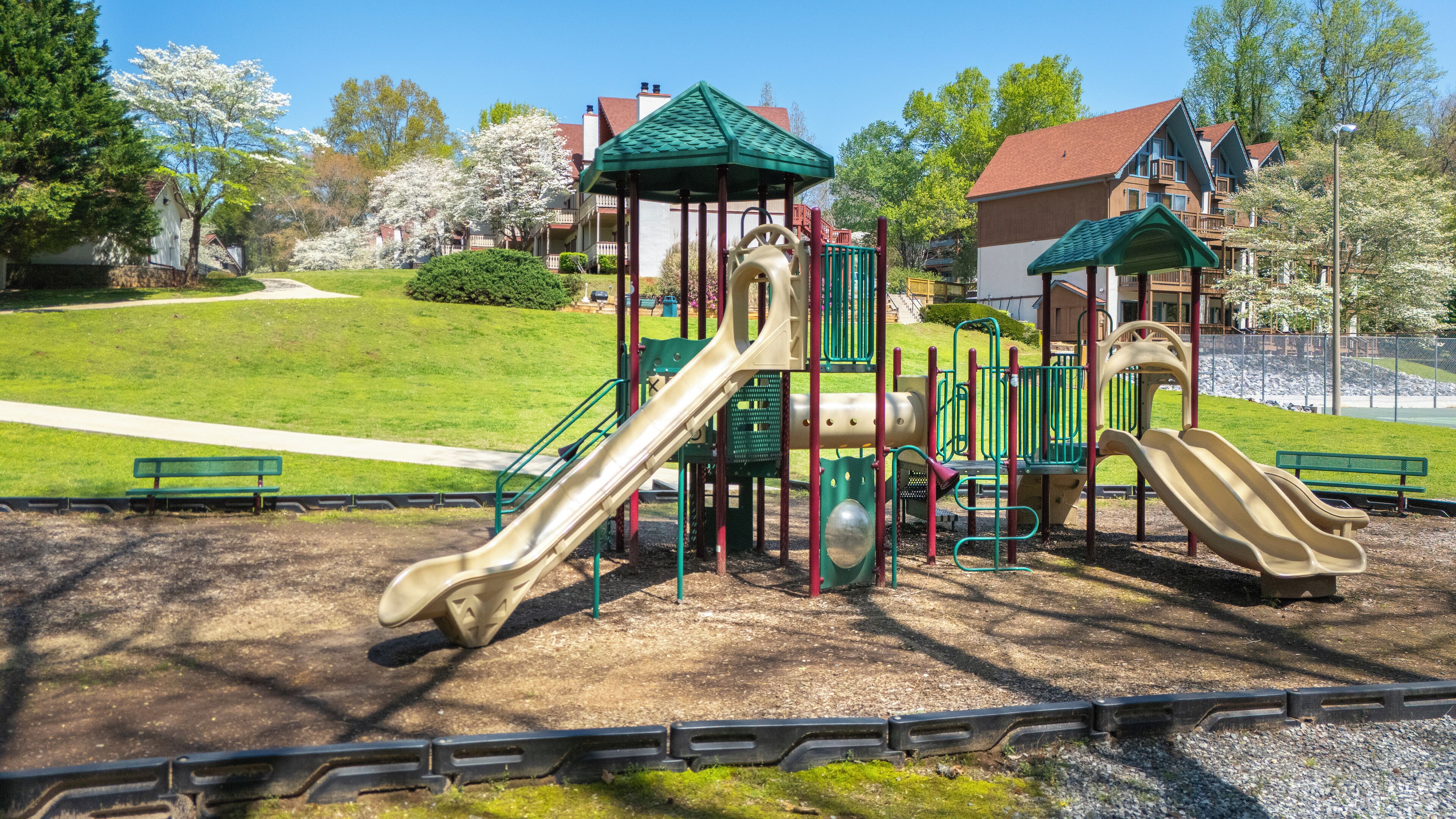 A playground with slides and climbing structures, surrounded by a park area with benches and a walking path.