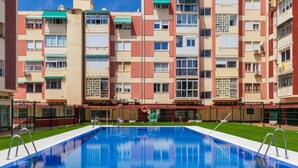 A swimming pool with a clear blue water surface, surrounded by a well-maintained lawn and a multi-story building with balconies and air conditioning units. - Holiday Home in Torremolinos by Playa Carihuela (Torremolinos)