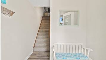 Una escalera que sube a un piso superior, un banco blanco con un cojín estampado y un espejo en la pared.