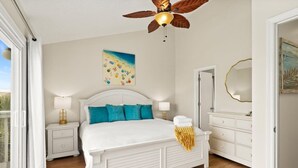 A bedroom with a white bed, teal pillows, a ceiling fan, a painting, and a white dresser.