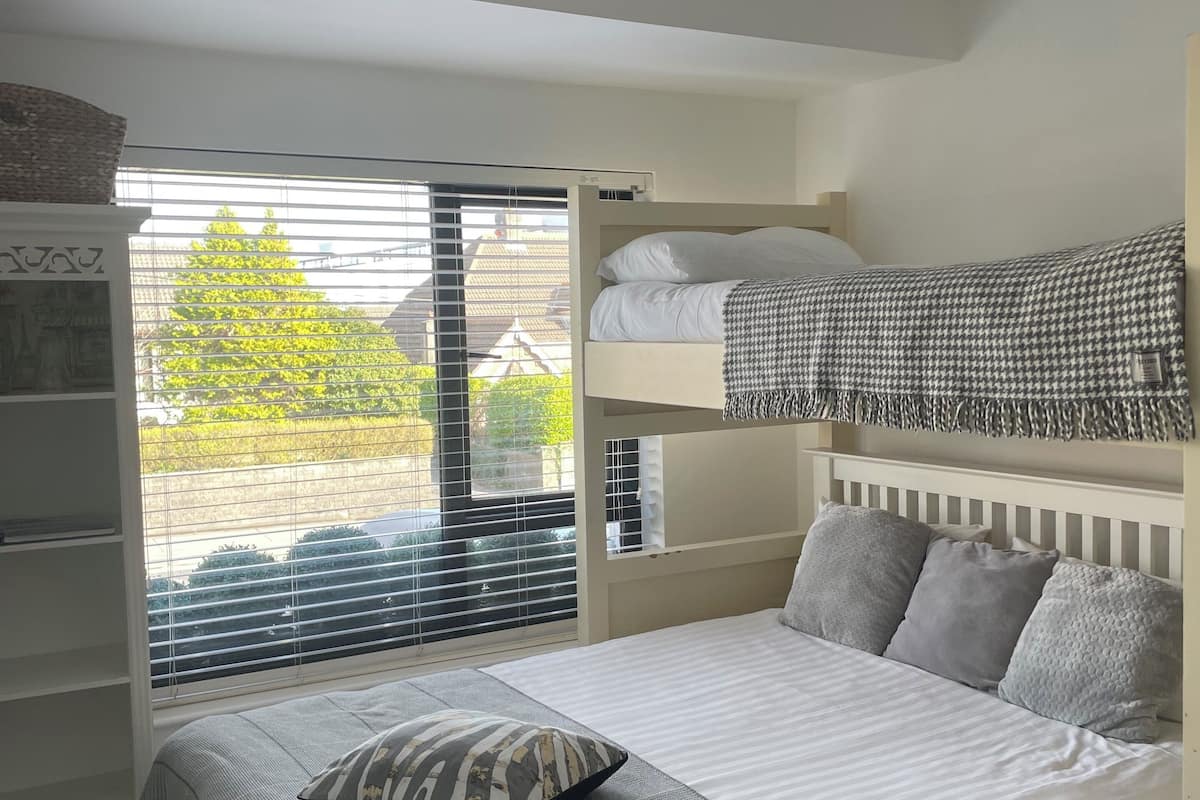 A bunk bed with a white headboard and grey bedding, a bedside table with a lamp, and a window with blinds overlooking a green area.
