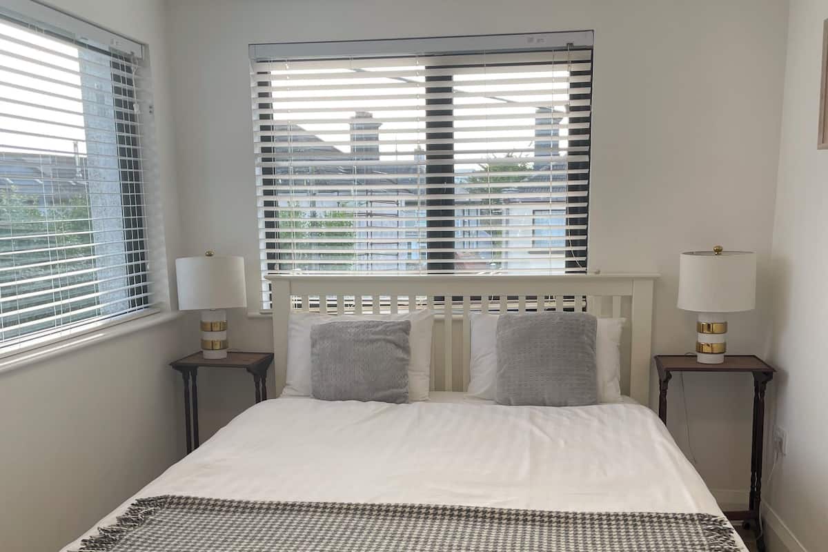 A neatly made bed with a houndstooth throw, two bedside lamps, and a window with blinds.