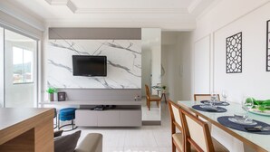 A modern dining area with a glass table, blue placemats, and a TV mounted on a marble wall. - Modern apt w/ Barbecue 600m From the Beach Com305 (Florianópolis)